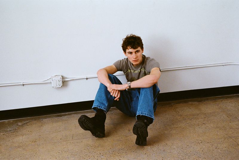 Sam Nivola sitting on a wood floor against a white wall wearing a grey t-shirt, blue jeans, and black boots.