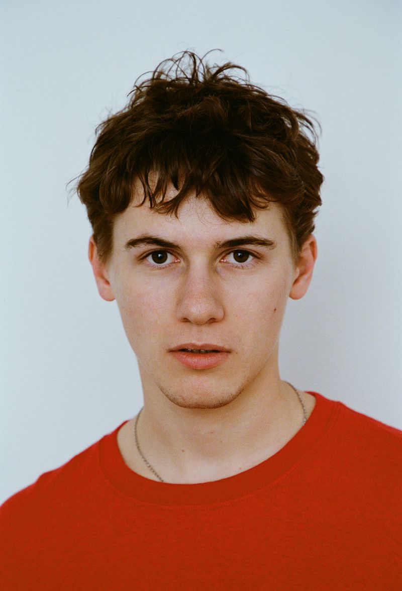 Portrait of a young adult male with dark hair, wearing a red t-shirt against a white background, looking directly at the camera.