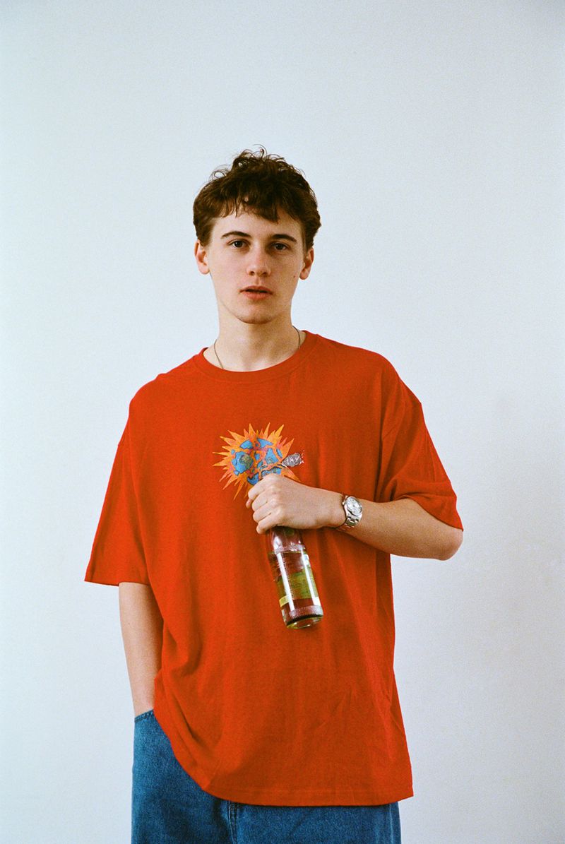 Young adult male poses indoors against a plain white backdrop, wearing a red t-shirt and jeans while holding a glass bottle.