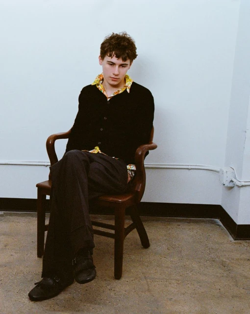 Young man sitting on wooden chair in black cardigan and patterned collared shirt against a plain white wall in a casual pose.