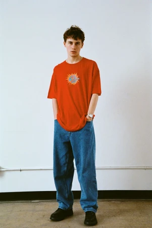 Sam Nivola wearing a red t-shirt and blue denim jeans standing against a plain white wall for HERO Magazine.