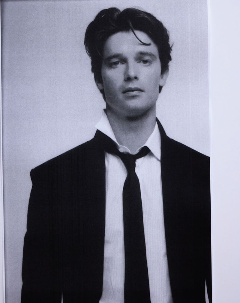 Portrait of Patrick Schwarzenegger wearing an unbuttoned white shirt and black tie with a dark suit jacket against a plain background