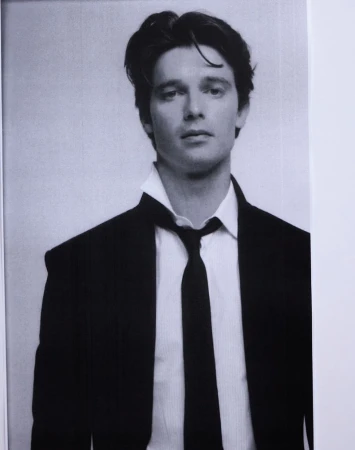 Portrait of Patrick Schwarzenegger wearing an unbuttoned white shirt and black tie with a dark suit jacket against a plain background