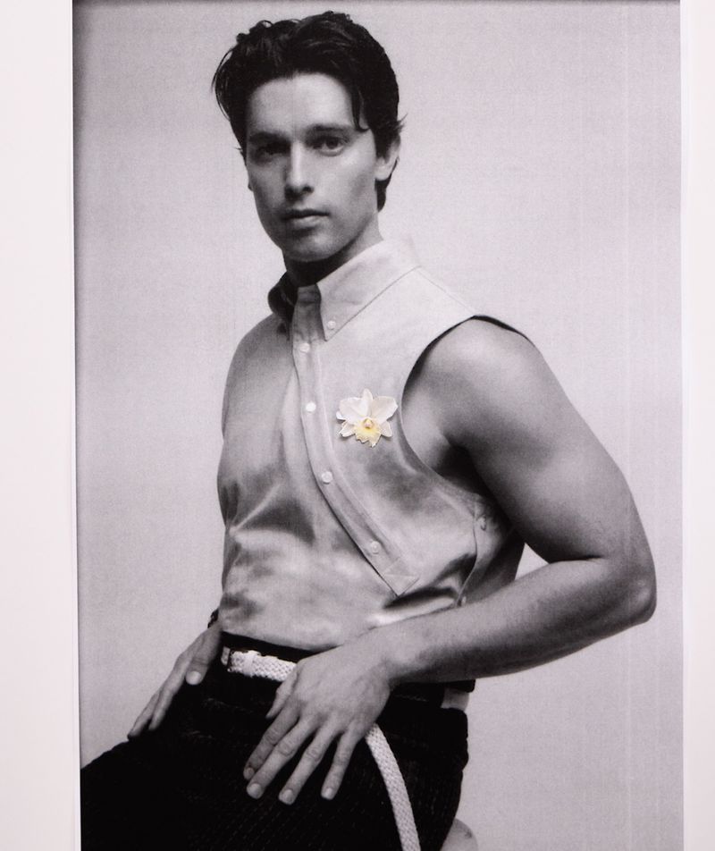 Portrait of a light-skinned man in a sleeveless shirt with a flower, posing indoors against a white background in monochrome style.