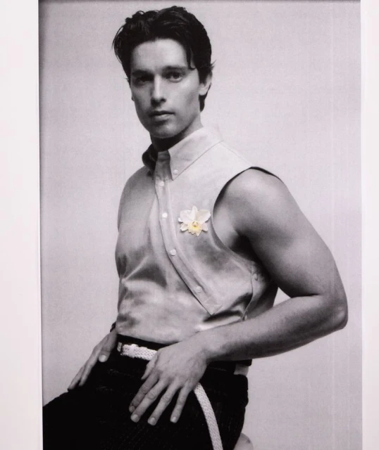 Portrait of a light-skinned man in a sleeveless shirt with a flower, posing indoors against a white background in monochrome style.