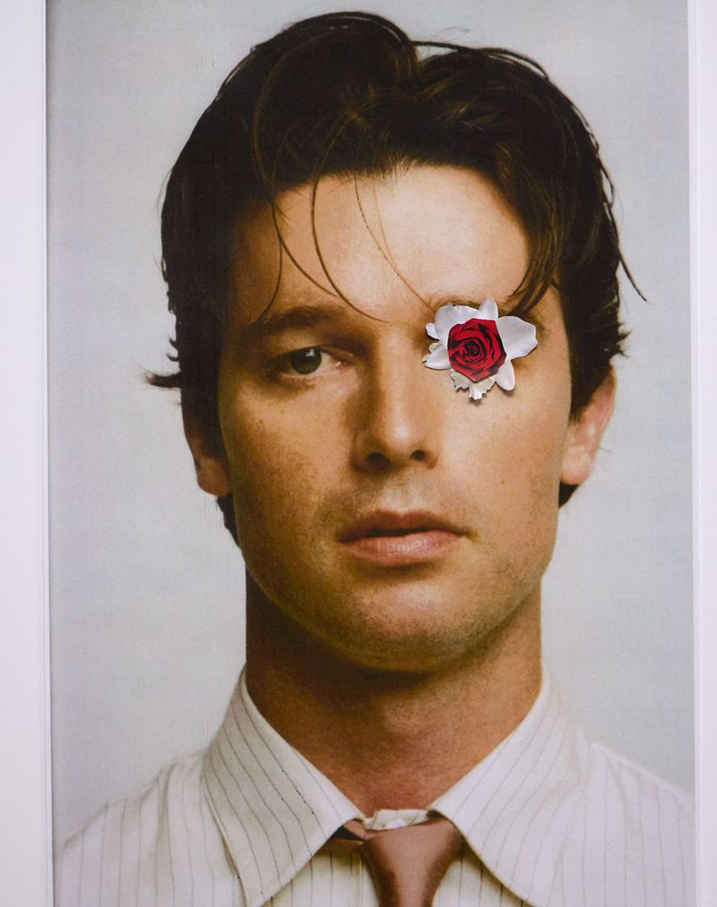 Close-up portrait of a dark-haired adult male in a striped shirt, with a red rose digitally composited over one eye.