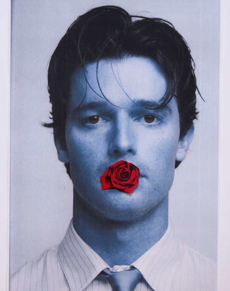 Young man in blue monochrome with a red rose covering his mouth, wearing a striped shirt and tie, creating a striking visual statement.