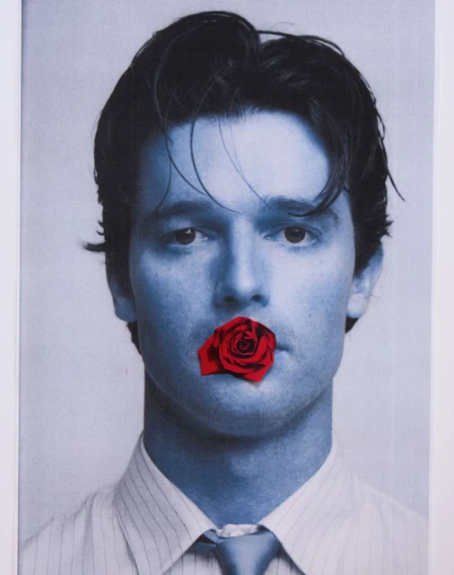 Young man in blue monochrome with a red rose covering his mouth, wearing a striped shirt and tie, creating a striking visual statement.
