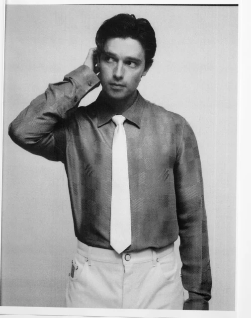 Monochrome portrait of an adult male wearing a patterned shirt and white tie, with hand raised to his head, against a plain backdrop.