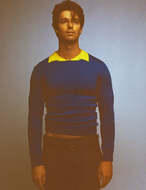 Studio shot of an adult male wearing a blue and yellow long-sleeved shirt and brown pants, posing against a neutral background.