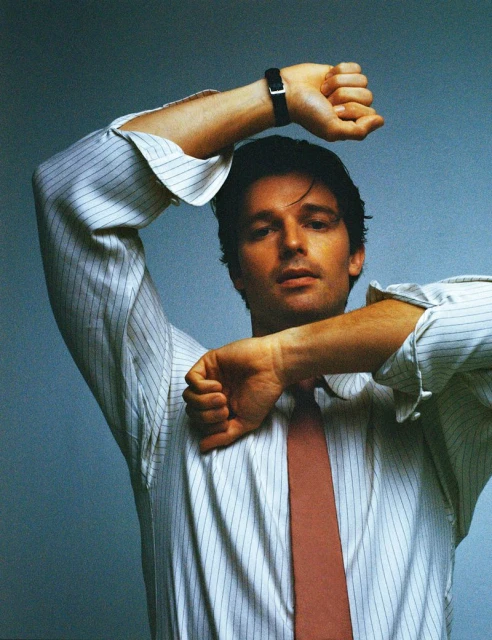 Portrait of a handsome adult male in a white striped shirt and tie, posed with his arms up against a grey background.