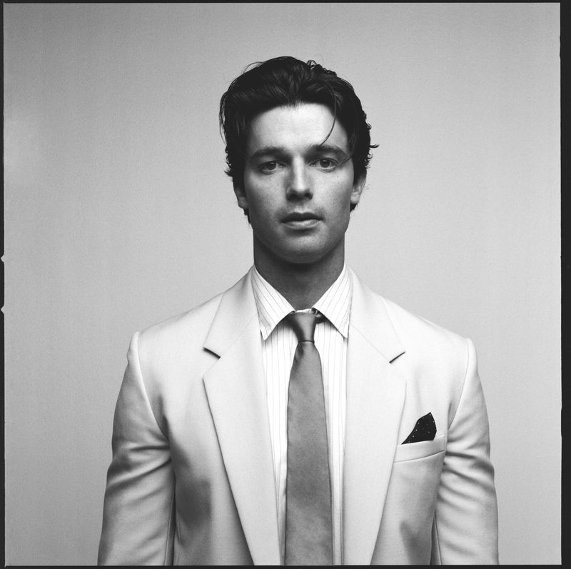 Monochrome portrait of Patrick Schwarzenegger wearing a light suit jacket, striped shirt, and tie against a plain backdrop.