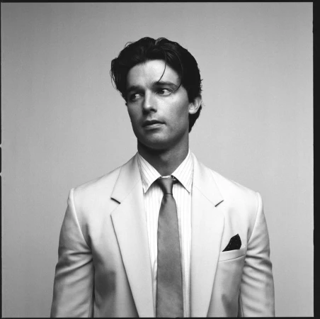 Headshot of an adult male with dark hair in a light-colored suit and tie, looking left against a neutral background in black and white.