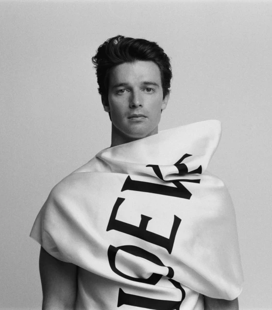 Portrait of a young adult male with dark hair wearing a large white fabric garment with black text, posing against a solid background.