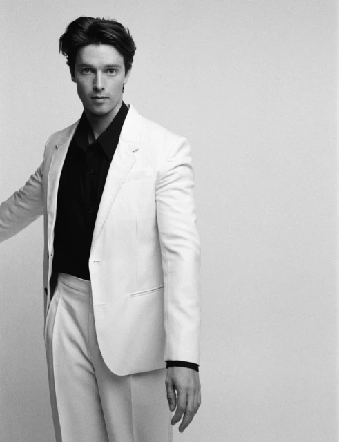 Monochrome studio portrait of an adult male in a white suit and black shirt against a neutral background, fashion and style.