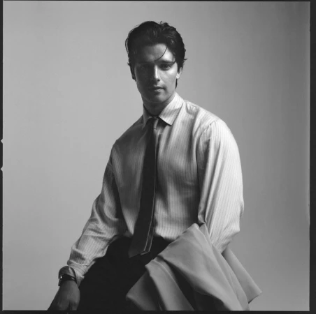 Portrait of Patrick Schwarzenegger wearing a striped shirt and tie against a plain studio background for HERO Magazine.
