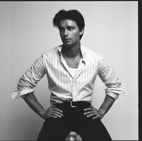 Monochrome portrait of Patrick Schwarzenegger wearing a striped shirt and dark trousers against a light backdrop.
