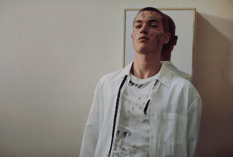 Portrait of a young adult man in a white shirt with face tattoos standing indoors in front of a mirror with a neutral expression.