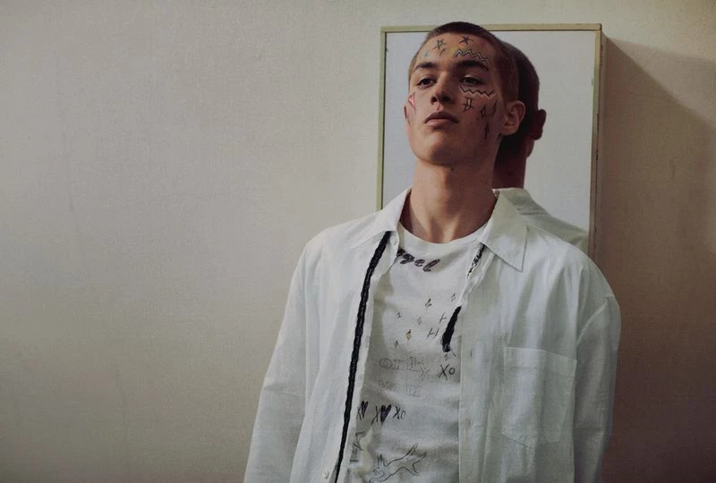 Portrait of a young adult man in a white shirt with face tattoos standing indoors in front of a mirror with a neutral expression.