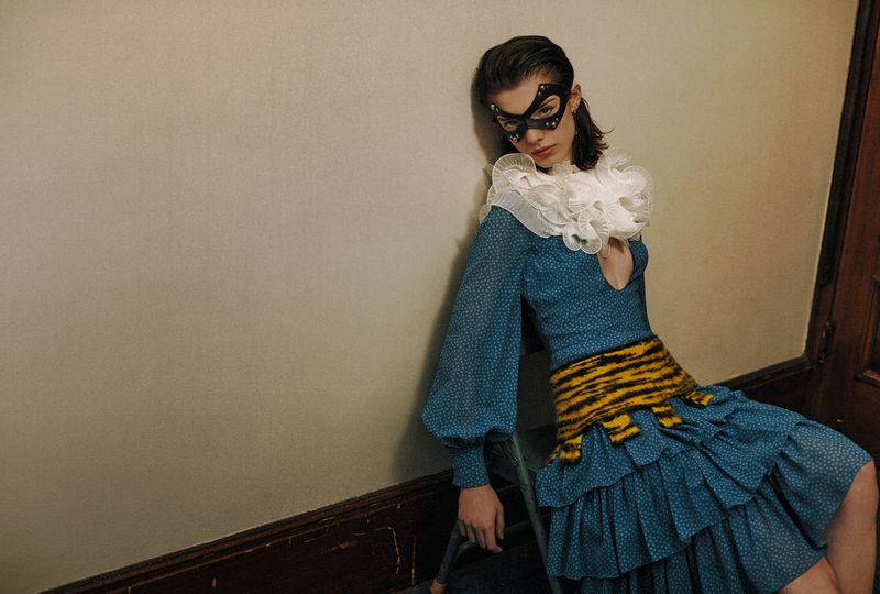 Woman in a blue dress with a white ruffled collar and black mask leans against a wall in an indoor setting, fashion photography.