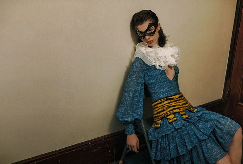 Woman in a blue dress with a white ruffled collar and black mask leans against a wall in an indoor setting, fashion photography.