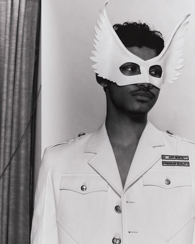 Monochrome portrait of a man wearing a wing mask and military-style jacket, standing in an indoor setting with a curtain.
