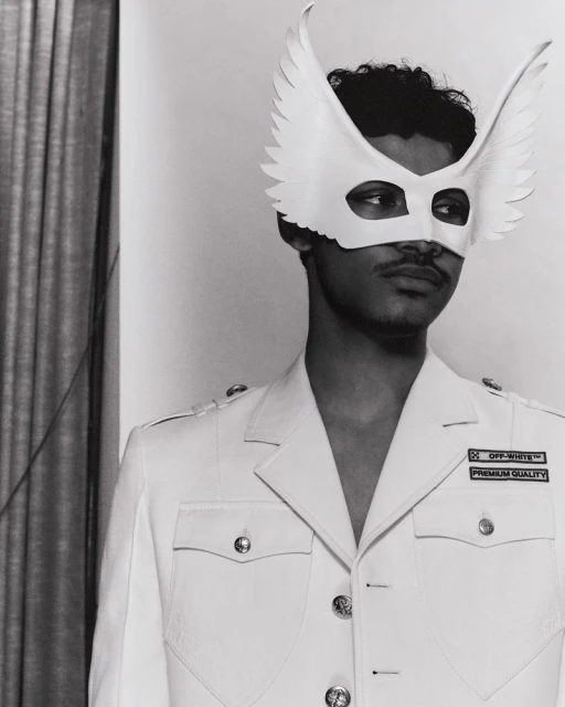 Monochrome portrait of a man wearing a wing mask and military-style jacket, standing in an indoor setting with a curtain.