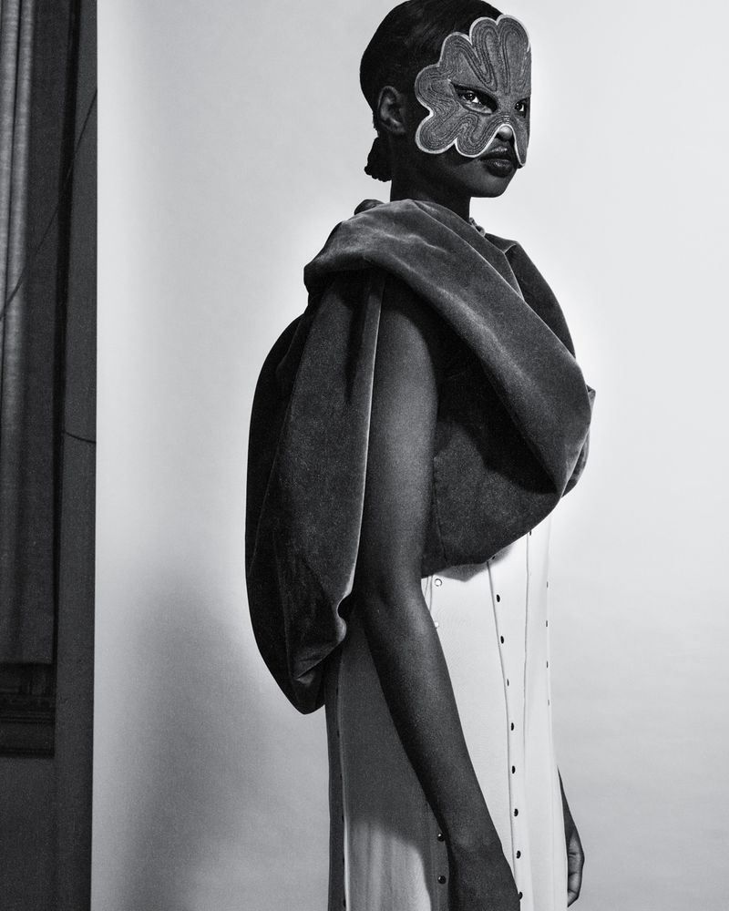 An adult female is posing indoors wearing a white dress with buttons and a large grey wrap. She is also wearing a flower mask.