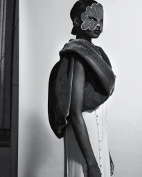 Black model wearing an embroidered mask and draped velvet garment over a white dress against a neutral backdrop