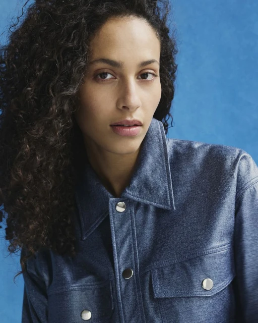 Close-up portrait of a woman with curly hair wearing a denim jacket against a solid blue background in a studio setting.