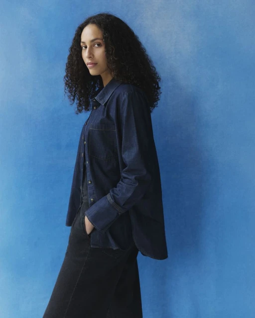 Portrait of a woman with curly dark hair wearing a denim shirt, standing in front of a bright blue background.