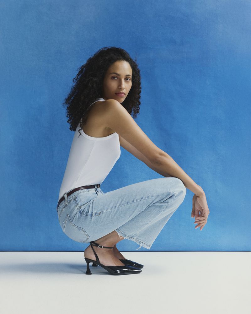 Model Anissa Bahkti wearing a white tank top and blue jeans squatting against a vibrant blue textured studio background.