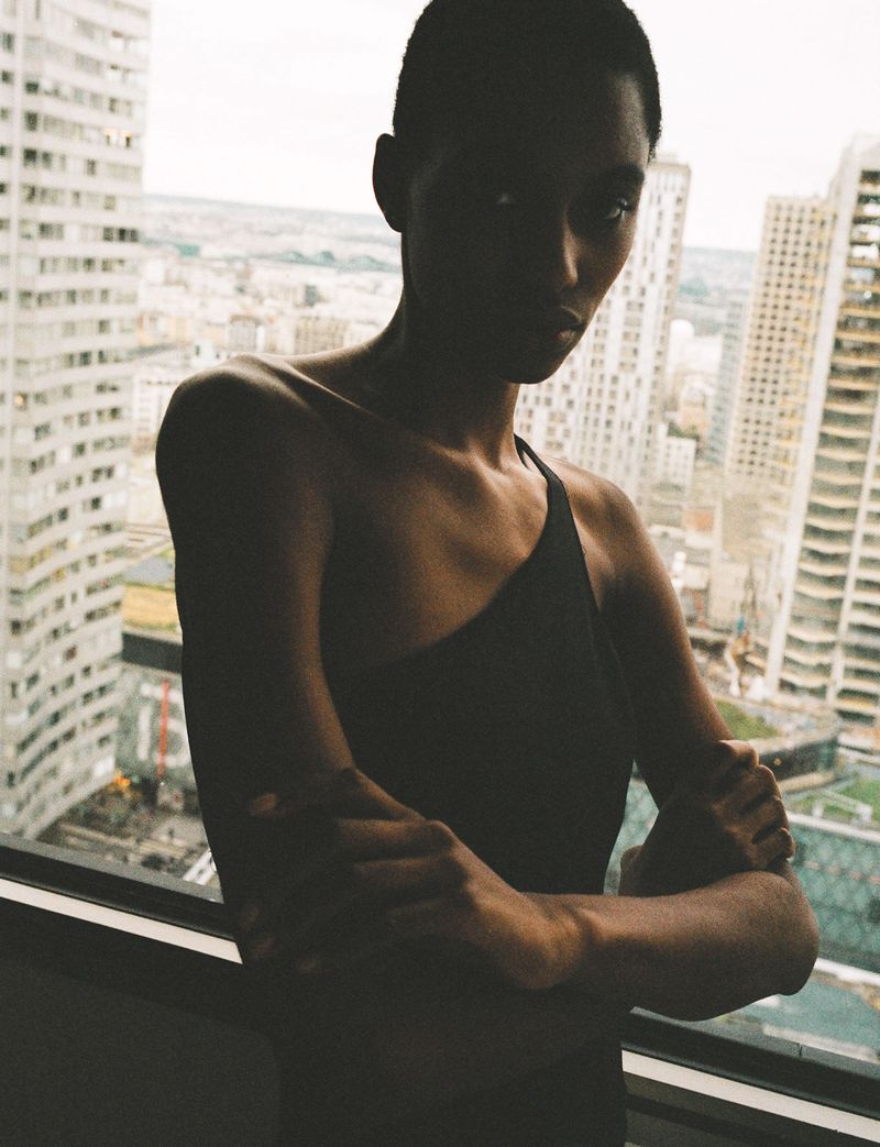 Indoor portrait of a Black adult woman with short hair, arms crossed, wearing a black top against a backdrop of high-rise buildings.