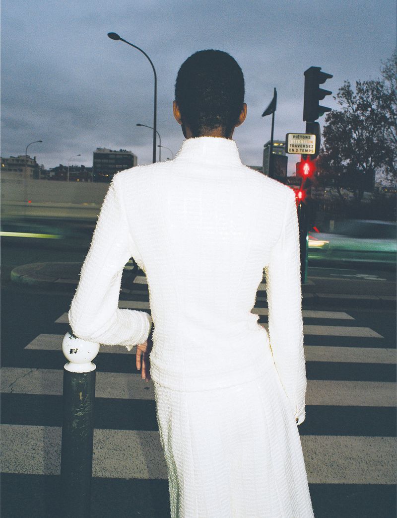 Rear view of an adult female in a white suit leaning on a bollard at a crosswalk during the evening in an urban setting.