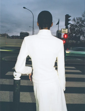 Rear view of an adult female in a white suit leaning on a bollard at a crosswalk during the evening in an urban setting.