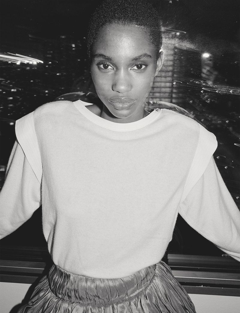 Monochrome portrait of a young Black woman in a light-colored sweater and shiny skirt, against a backdrop of blurred city lights.