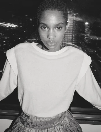 Monochrome portrait of a young Black woman in a light-colored sweater and shiny skirt, against a backdrop of blurred city lights.