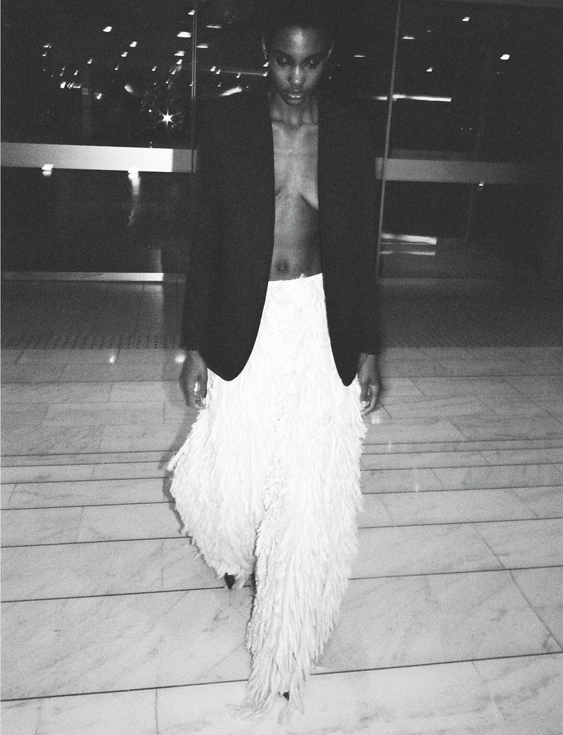 A slender woman with an updo, wearing a black blazer and fringed trousers, descends marble stairs in a dimly lit interior.
