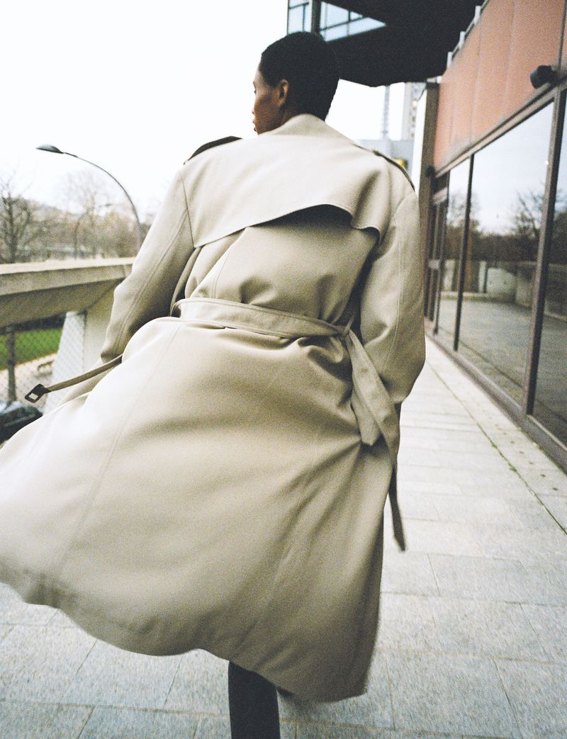 A woman in a flowing trench coat walks along a paved walkway near a modern building on an overcast day, fashion photography, outdoor setting.