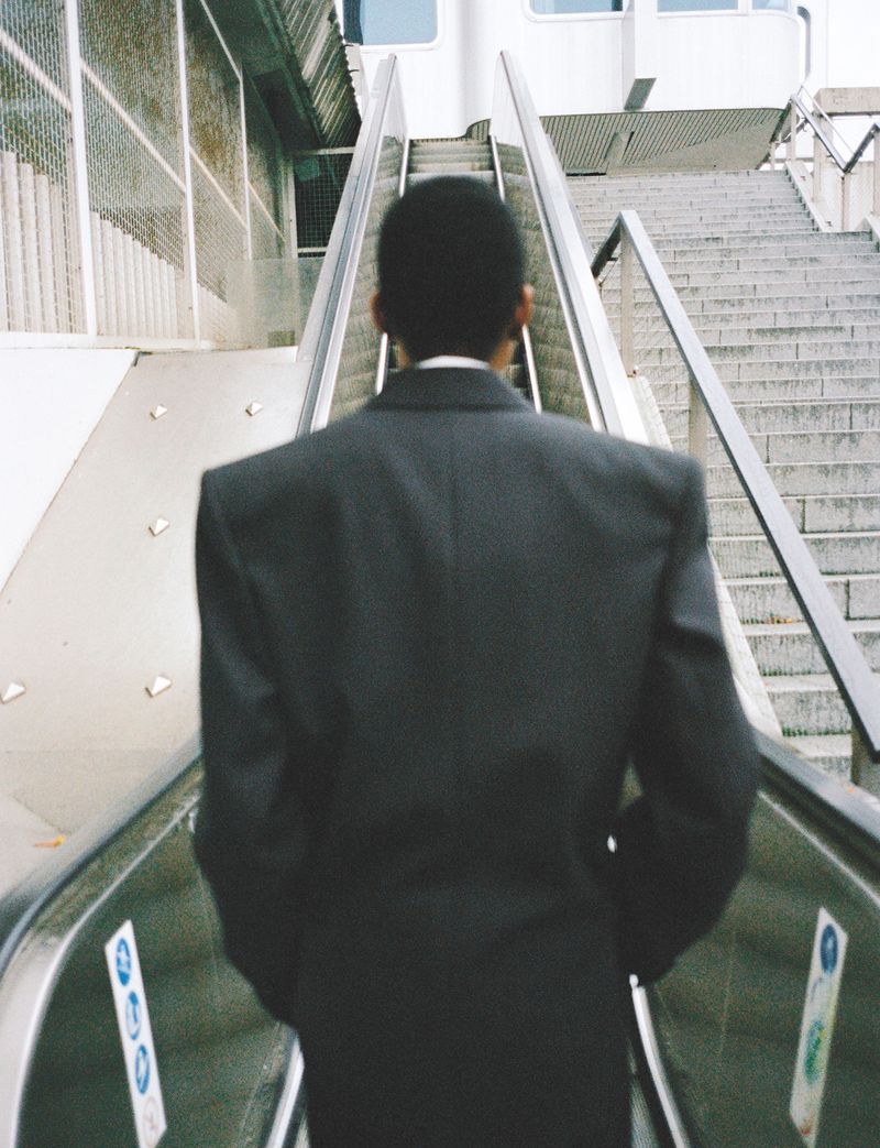 Back view of a Black adult male wearing a grey suit riding on an escalator toward stairs in an urban environment, daytime.