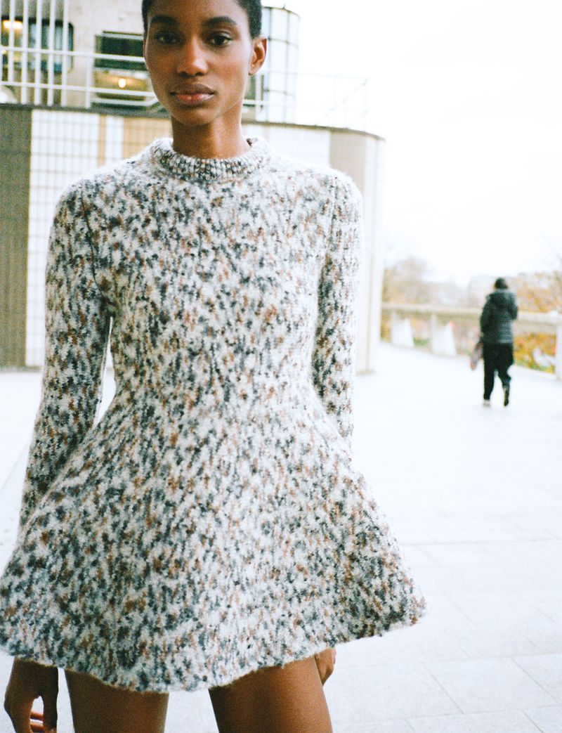 A fashionable young Black woman in a knit dress stands outdoors with blurred background figure, showcasing style and urban aesthetic.