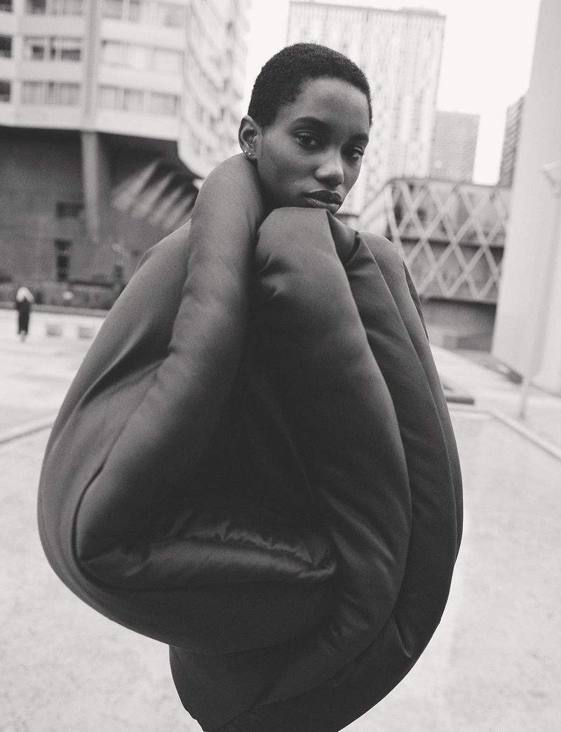 Black and white fashion shot of a woman with short hair wearing an oversized padded jacket against a neutral city background.