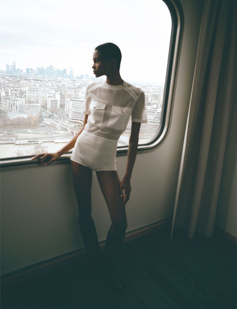 A slim, elegant Black woman in white outfit poses by a large window, with a cityscape in the background under an overcast sky.