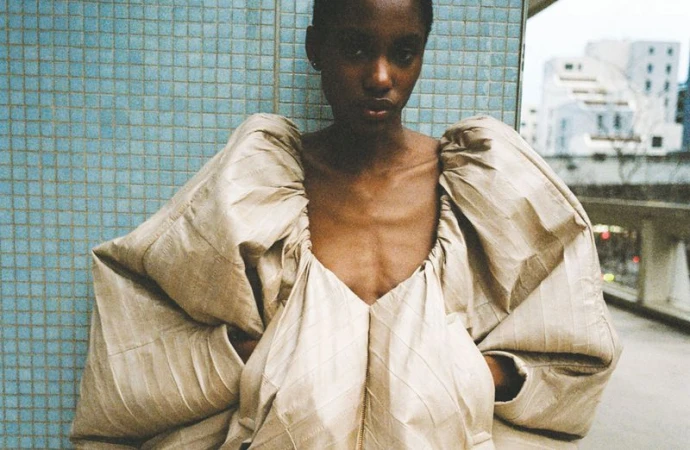 Fashionable adult woman in an avant-garde dress posing against a blue tiled wall in an urban setting, with buildings in the background.