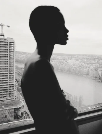 Silhouette of a person with short hair looking out a high-rise window overlooking a city and river.