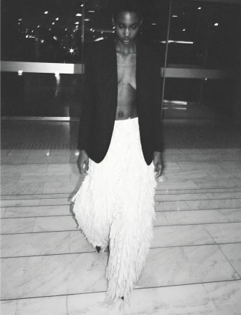 Model Tara Falla in a black blazer and white fringed skirt walking across a tiled floor for Muse Magazine.