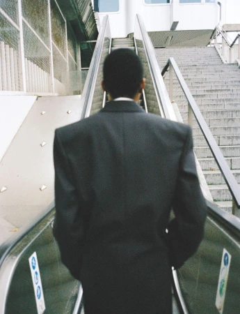 Rear view of a person wearing a dark oversized blazer riding an outdoor escalator alongside concrete stairs.