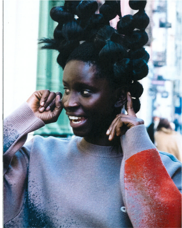 Portrait of a dark-skinned woman with elaborate hair, wearing a grey sweater with colorful accents, set in an urban environment.