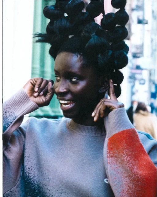 Portrait of a dark-skinned woman with elaborate hair, wearing a grey sweater with colorful accents, set in an urban environment.