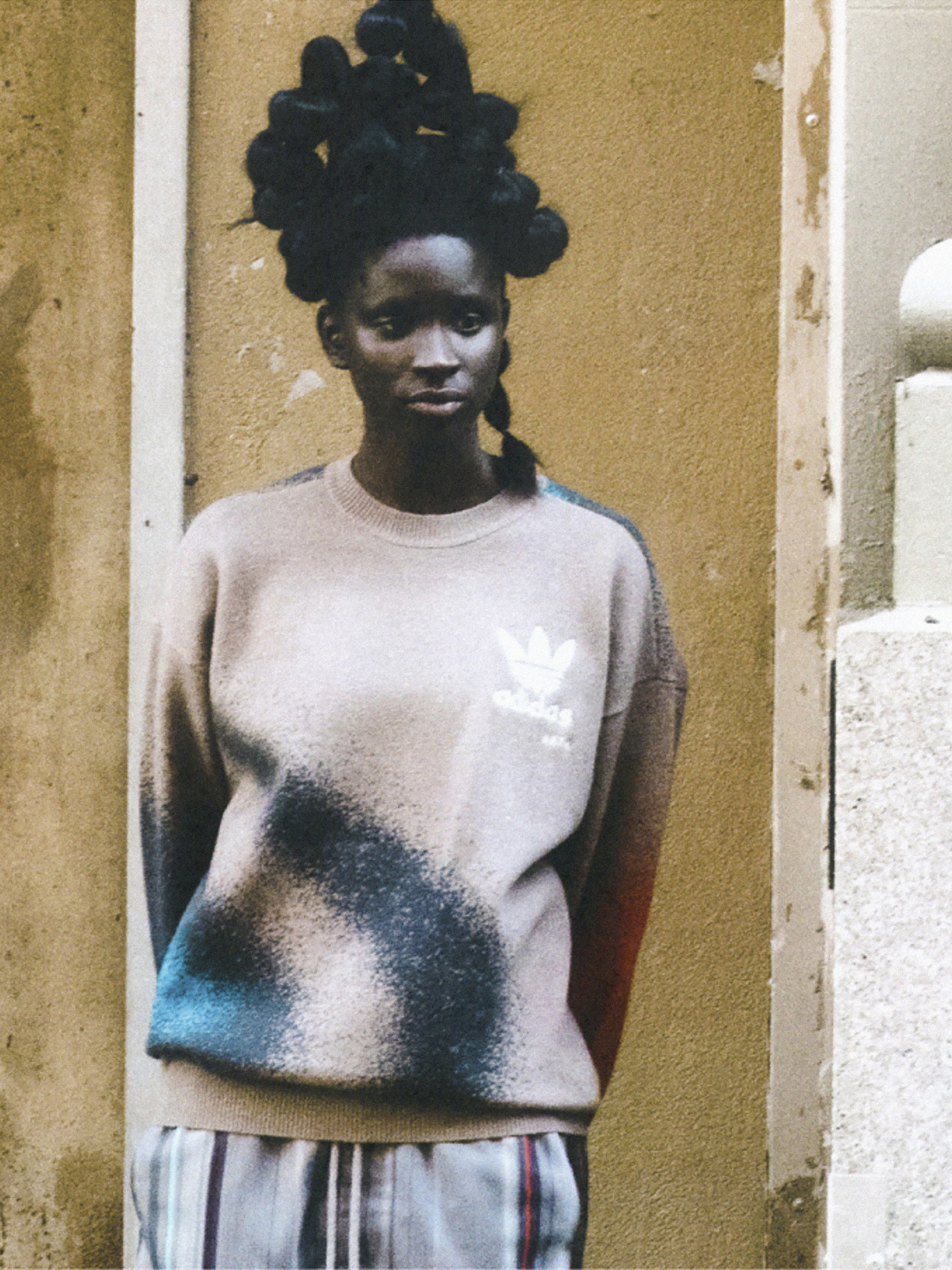 Woman in Adidas sweatshirt poses against a textured wall. She has a unique hairstyle, fair skin, and stylish striped pants.